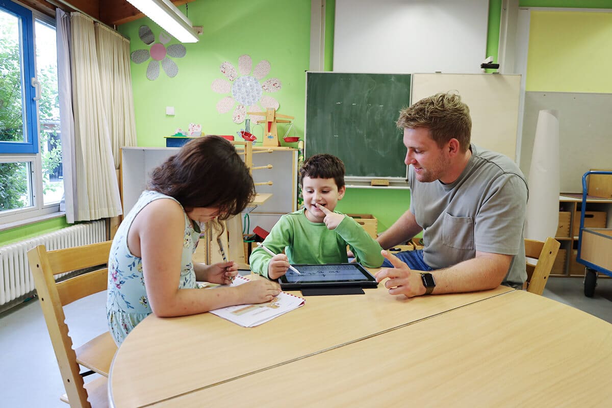 Zwei Kinder und ein Lehrer sitzen gemeinsam am Tisch und arbeiten sowohl im Heft als auch mit einem Tablet