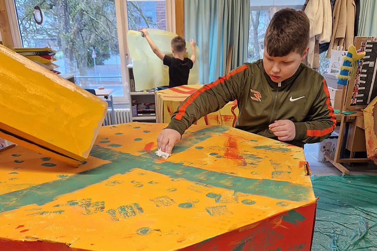 Junge gestaltet mit einem Schwamm eine große, gelb-orange bemalte Pappstruktur