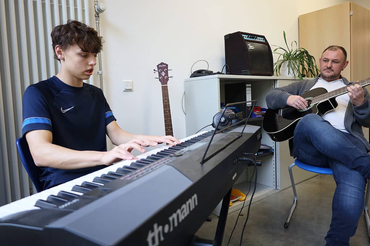 Zwei Personen sitzen in einem Raum, einer spielt Keyboard, der andere Gitarre
