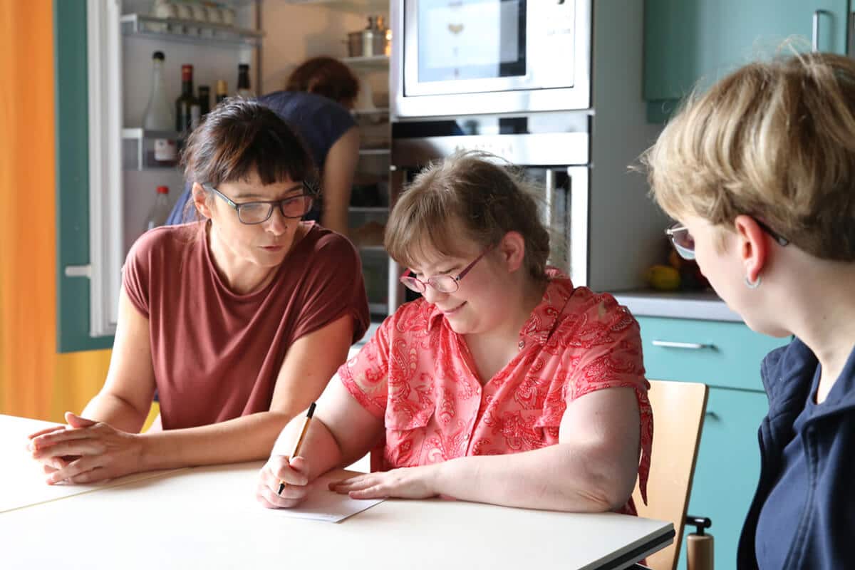 Drei Personen sitzen an einem Tisch in einer Küche, eine Person schreibt auf Papier, die anderen beiden schauen zu